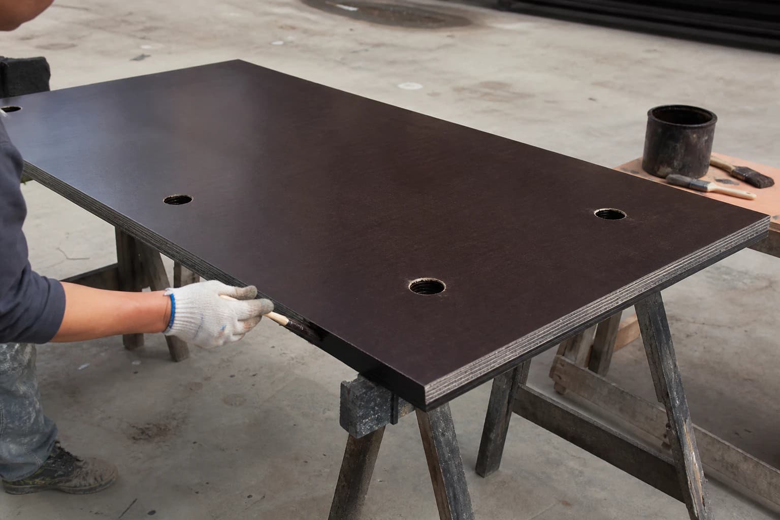 Worker applying waterproof edge-sealing paint only to a freshly cut film-faced plywood edge on aligned trestles