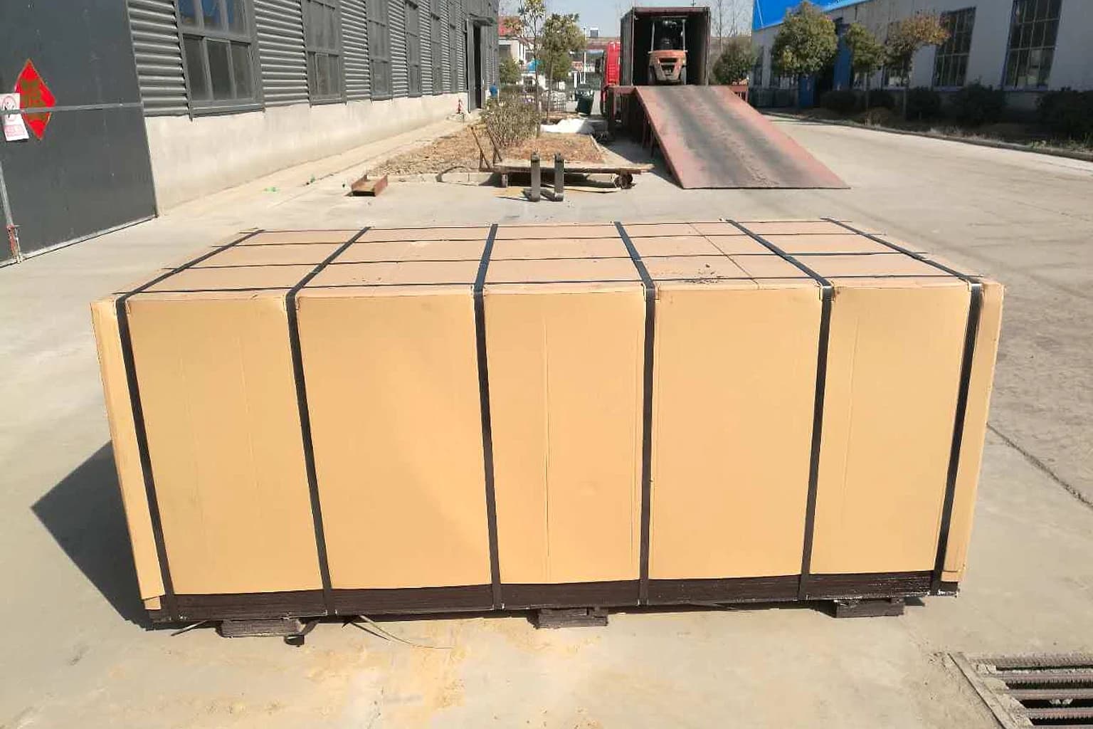 Cardboard-wrapped plywood bundle with continuous black straps on bearers beside a truck loading ramp