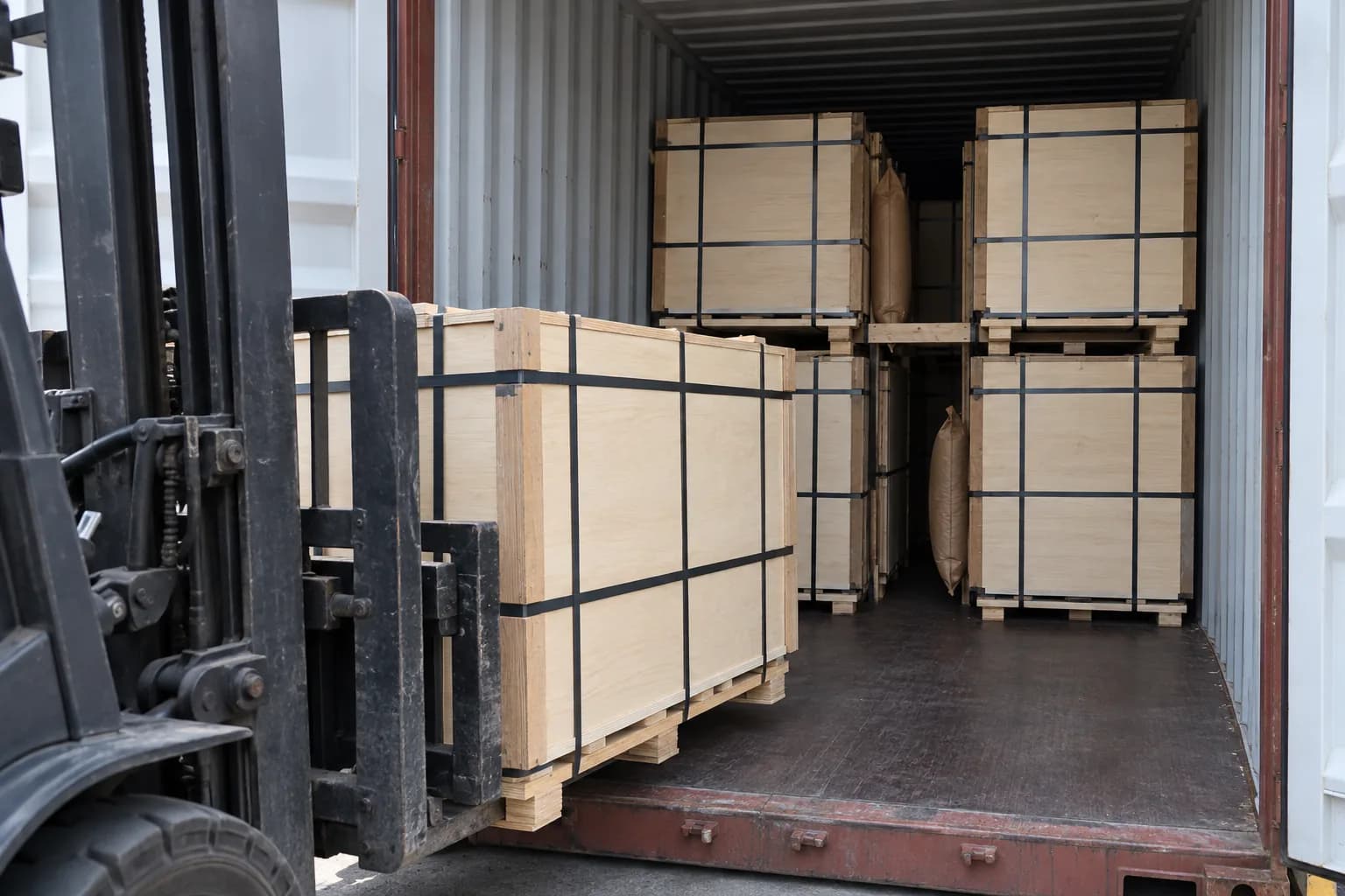 Industrial forklift placing a long strapped flat plywood pallet into a freight container with a spotter remaining clear of the movement zone